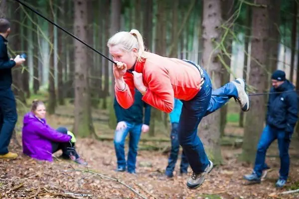 Frau überquert selbstgebaute Seilbrücke im Wald, während andere zuschauen