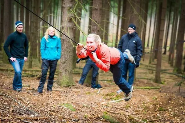 Frau überquert selbstgebaute Seilbrücke im Wald, Zuschauer beobachten