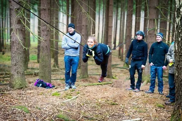 Frau überquert selbstgebaute Seilbrücke zwischen Bäumen, Zuschauer beobachten