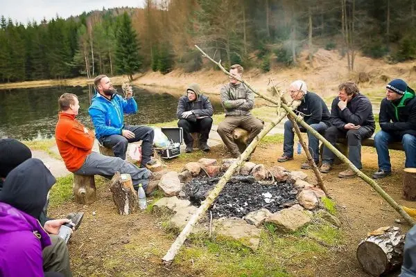 Gruppe sitzt um ein kleines Bodenfeuer, einer erklärt etwas mit einem Stock