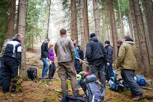 Gruppe von Personen steht im Wald um eine Holzstange, Rucksäcke liegen am Boden