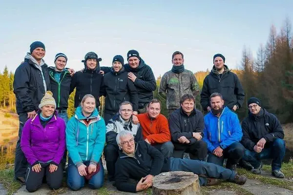 Gruppenszenario im Freien, Teilnehmer in Outdoor-Bekleidung stehen und sitzen um einen Baumstumpf
