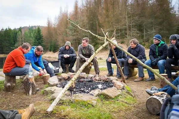Lagerfeuer mit Steinen umgeben, mehrere Personen sitzen auf Baumstümpfen