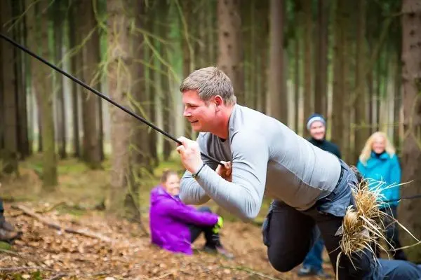 Mann überquert selbstgebaute Seilbrücke im Wald, andere Personen im Hintergrund