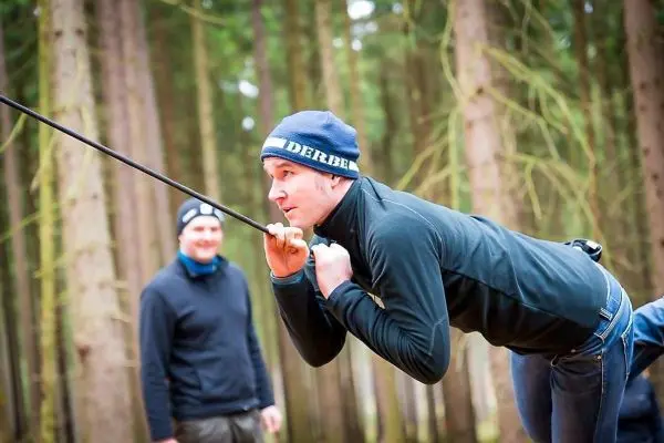 Mann überquert selbstgebaute Seilbrücke im Wald, während ein anderer zuschaut