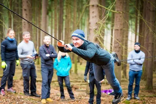 Mann überquert selbstgebaute Seilbrücke im Wald, Zuschauer beobachten