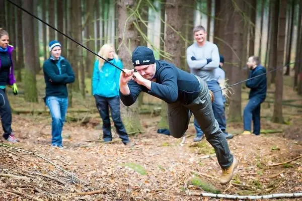 Mann überquert selbstgebaute Seilbrücke im Wald, Zuschauer beobachten