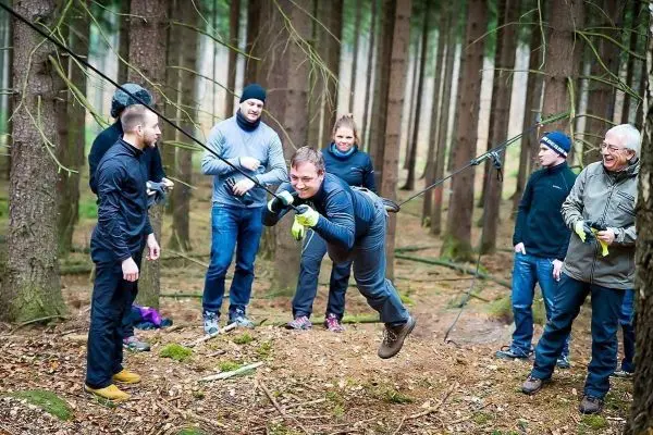 Mann überquert selbstgebaute Seilbrücke zwischen Bäumen, Zuschauer beobachten