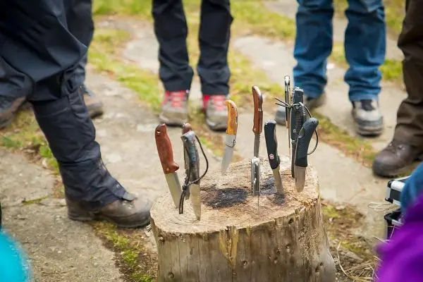 Mehrere Messer stecken in einem Baumstumpf, umgeben von Personen in Outdoor-Bekleidung