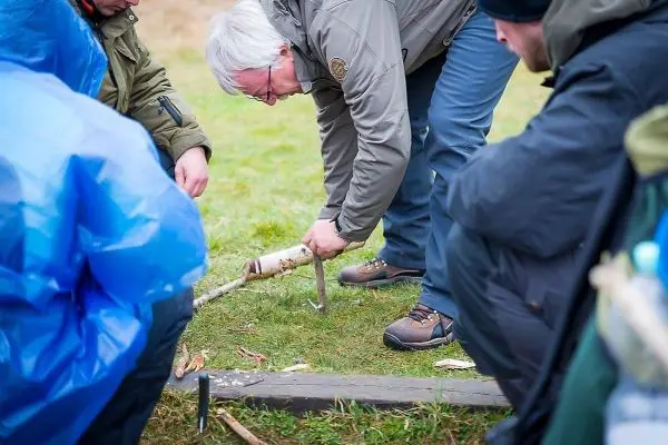 Messer bearbeitet Holzstück auf dem Boden, während andere Personen beobachten