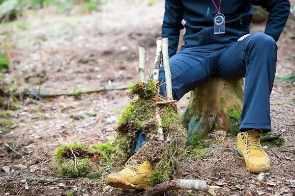 Person sitzt auf einem Baumstumpf neben einem selbstgebauten Beinprothese aus Ästen und Moos beim Survival Training