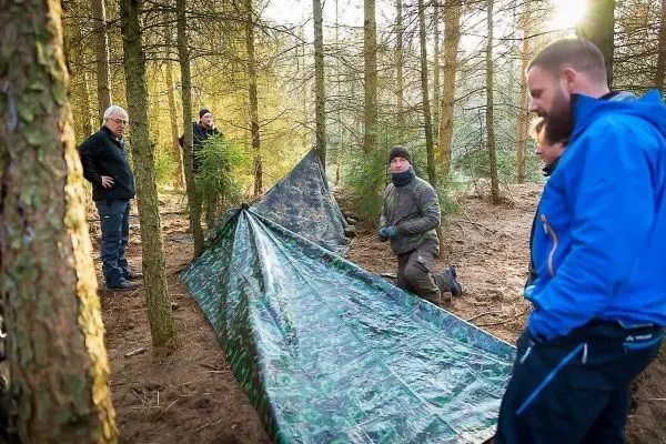 Tarp-Notunterkunft wird zwischen Bäumen aufgebaut, mehrere Personen beobachten den Aufbau