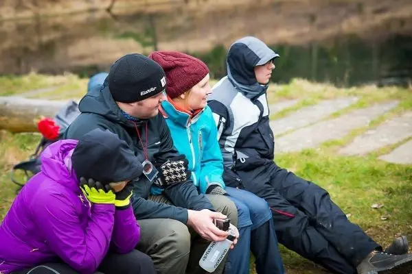 Vier Personen sitzen auf einer Bank am Ufer eines Gewässers, einige tragen warme Kleidung