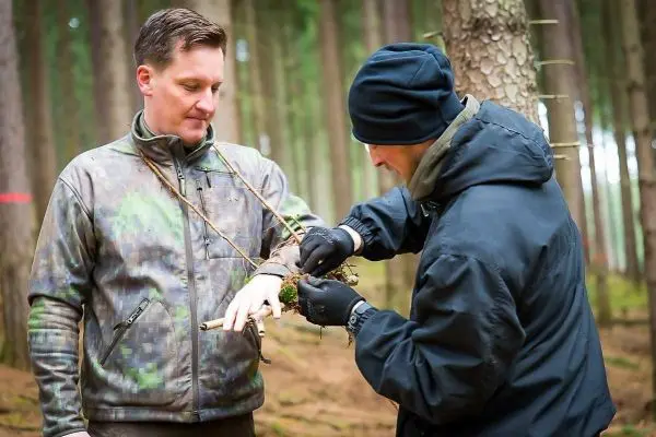 Zwei Personen binden improvisierte Materialien um ein Handgelenk im Wald