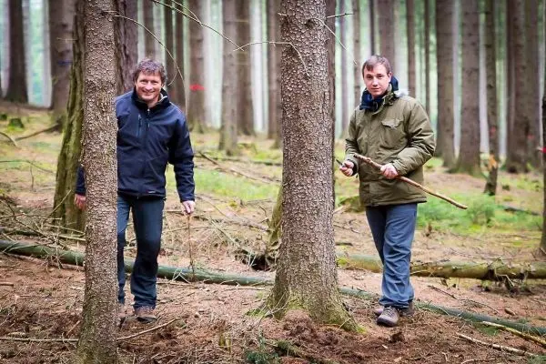 Zwei Personen gehen zwischen Bäumen im Wald, einer hält einen Stock in der Hand