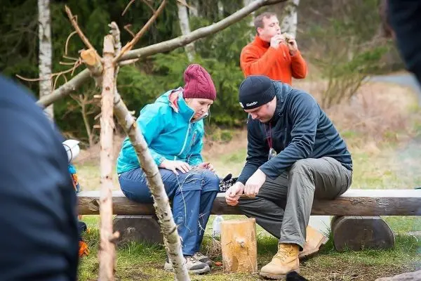 Zwei Personen sitzen auf einer Bank und bearbeiten Holz mit einem Messer