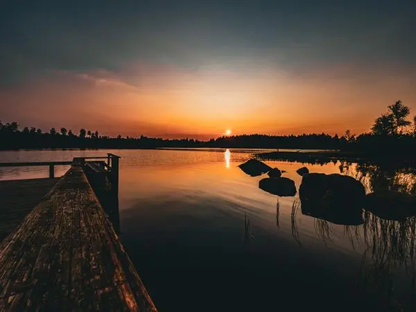 Holzsteg führt zu ruhigem Wasser bei Sonnenuntergang, umgeben von Bäumen und Felsen