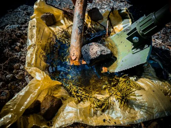 Holzstück steht in einer gelben Plane mit Wasser und Steinen, daneben eine Schaufel