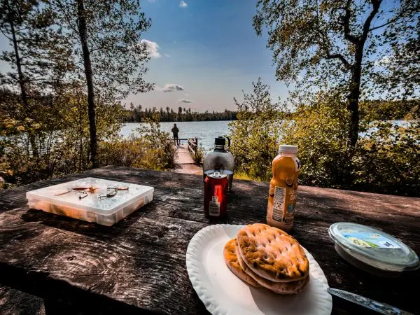 Holztisch mit Snacks und Getränken, im Hintergrund ein Angler am Wasser