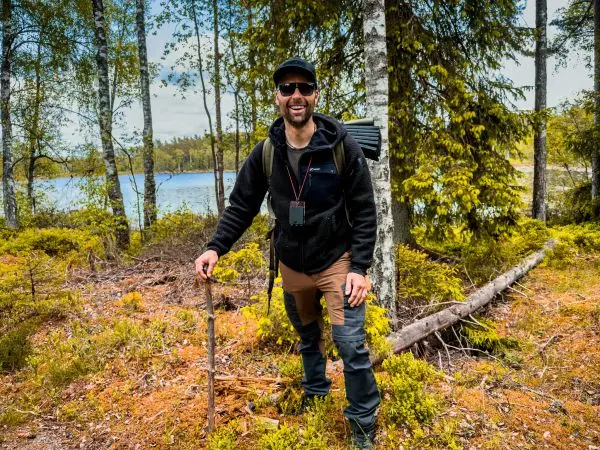 Mann mit Wanderstock steht im Wald, im Hintergrund ein Gewässer und Bäume
