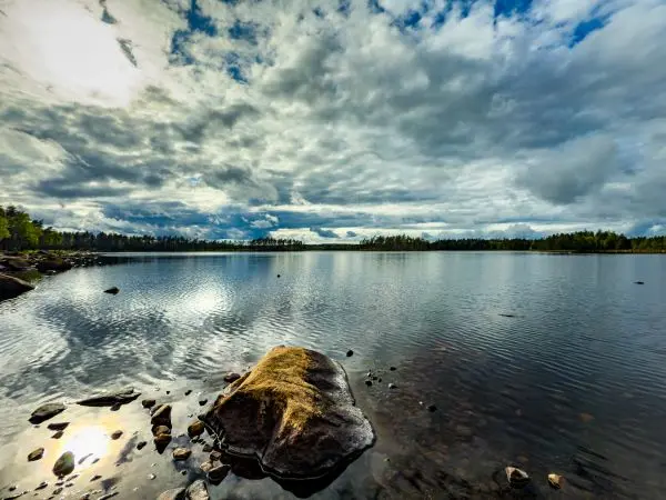Stillgewässer mit spiegelndem Wasser und Wolken am Himmel, umgeben von Bäumen und Steinen
