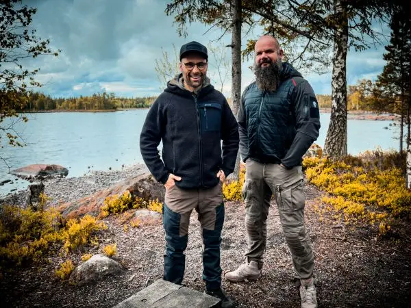 Zwei Männer stehen am Ufer eines Sees, umgeben von Bäumen und Steinen