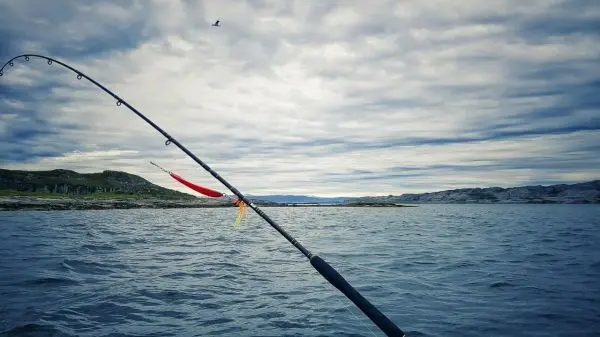 Angelrute mit Köder über Wasser, im Hintergrund Wolken und Küstenlinie sichtbar