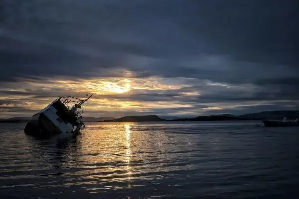 Ein gesunkenes Boot steht schräg im Wasser, während die Sonne am Horizont aufgeht