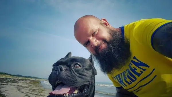 Ein Mann mit Bart und gelbem Shirt posiert mit einem schwarzen Hund am Strand