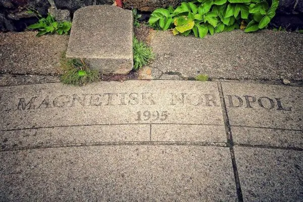 Granitplatte mit eingravierter Inschrift „MAGNETISK NORDPOL 1995“ und umgebender Vegetation