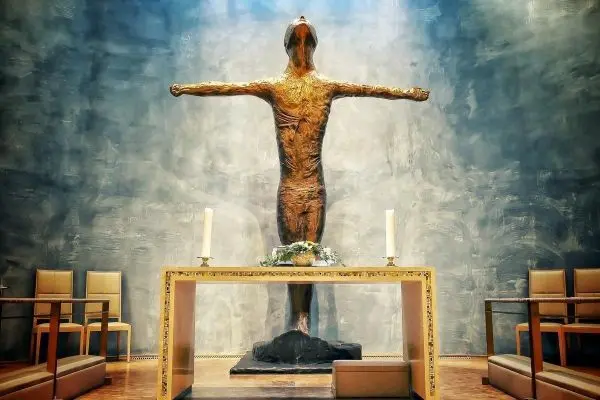 Skulptur eines menschlichen Körpers mit ausgebreiteten Armen vor einem Altar
