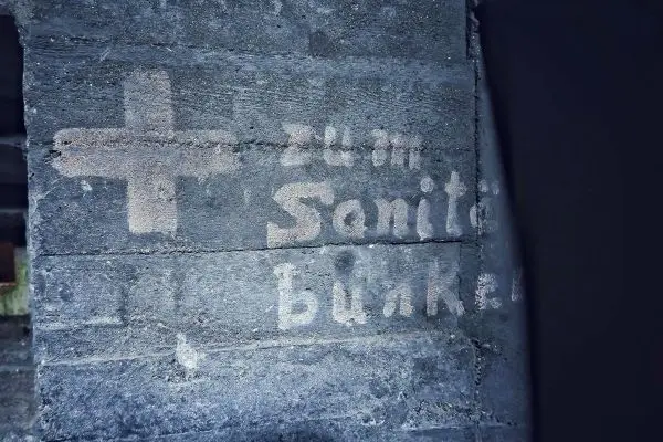 Wand mit verblasster Schrift und einem Kreuz, Hinweis auf Sanitärbunker