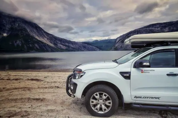Weißer Geländewagen parkt am Ufer eines Sees, im Hintergrund Berge und Wolken