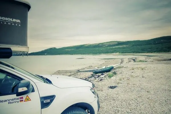 Weißes Fahrzeug steht am Ufer eines Gewässers mit einem Boot im Hintergrund