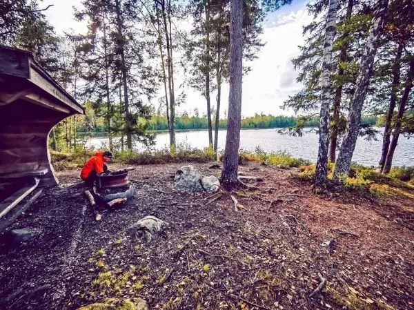 Person in orange Jacke bereitet Feuerstelle am Ufer eines Sees vor
