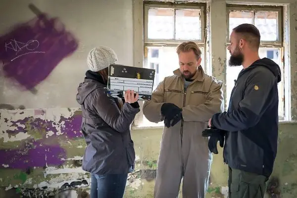 Drei Personen in einem Raum mit abblätternder Farbe, eine Person hält eine Klappe