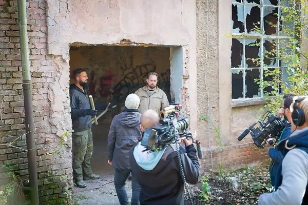 Drei Personen stehen im Eingang eines alten Gebäudes, Kamerateams filmen die Szene