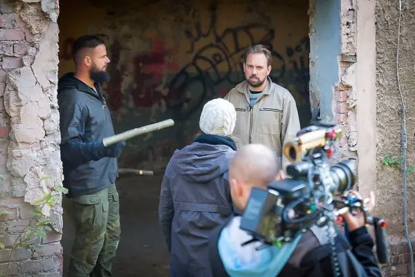 Drei Personen stehen in einem verfallenen Raum, eine Kamera filmt die Szene
