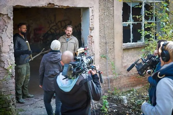 Drei Personen stehen vor einem verfallenen Gebäude, Kameras zeichnen die Szene auf