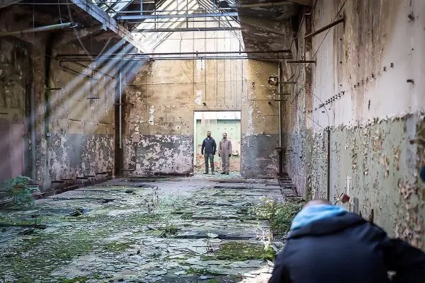 Verfallenes Gebäude mit Pflanzenbewuchs und Lichtstrahlen, zwei Personen stehen im Hintergrund