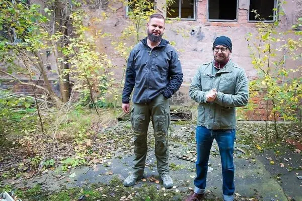 Zwei Männer stehen in einer verwilderten Umgebung mit Pflanzen und einer alten Mauer