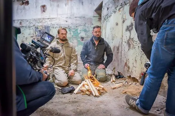 Zwei Personen sitzen vor einem Feuer in einem Raum mit beschädigten Wänden