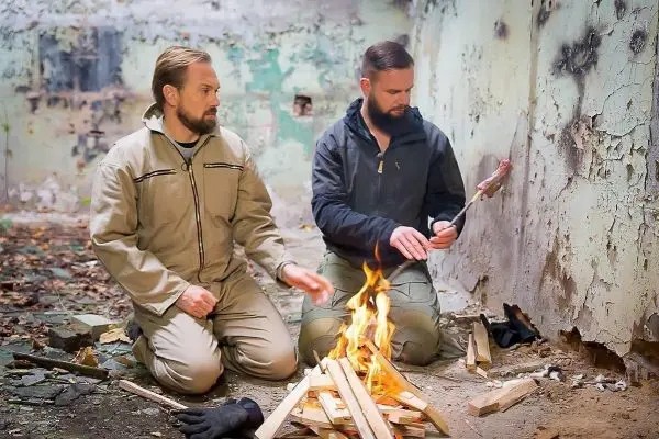 Zwei Personen sitzen vor einem Feuer und garen Fleischspieße über den Flammen