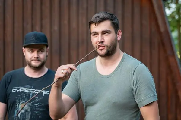 Mann hält einen Stock im Mund und spricht, während ein anderer Mann im Hintergrund steht