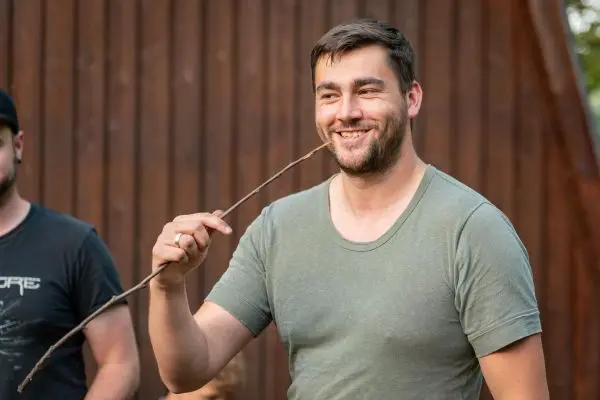 Mann hält einen Stock und lächelt vor einer Holzwand