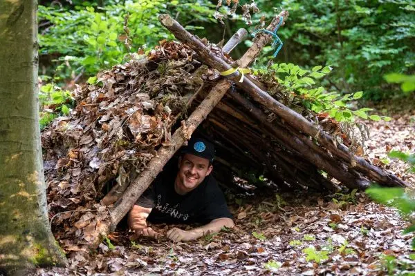 Mann liegt in einer Laubhütte aus Ästen und Blättern im Wald