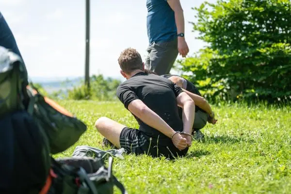 Person sitzt mit gefesselten Händen auf dem Gras, während eine andere Person in der Nähe steht