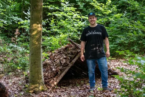 Person steht neben einer selbstgebauten Unterkunft im Wald