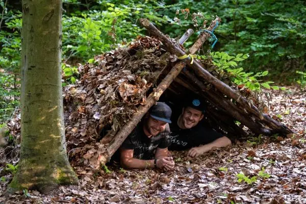 Zwei Personen liegen in einer Laubhütte aus Ästen und Blättern