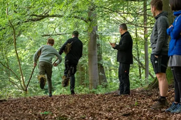 Zwei Personen mit Tarnmaterial laufen durch den Wald, während andere zuschauen